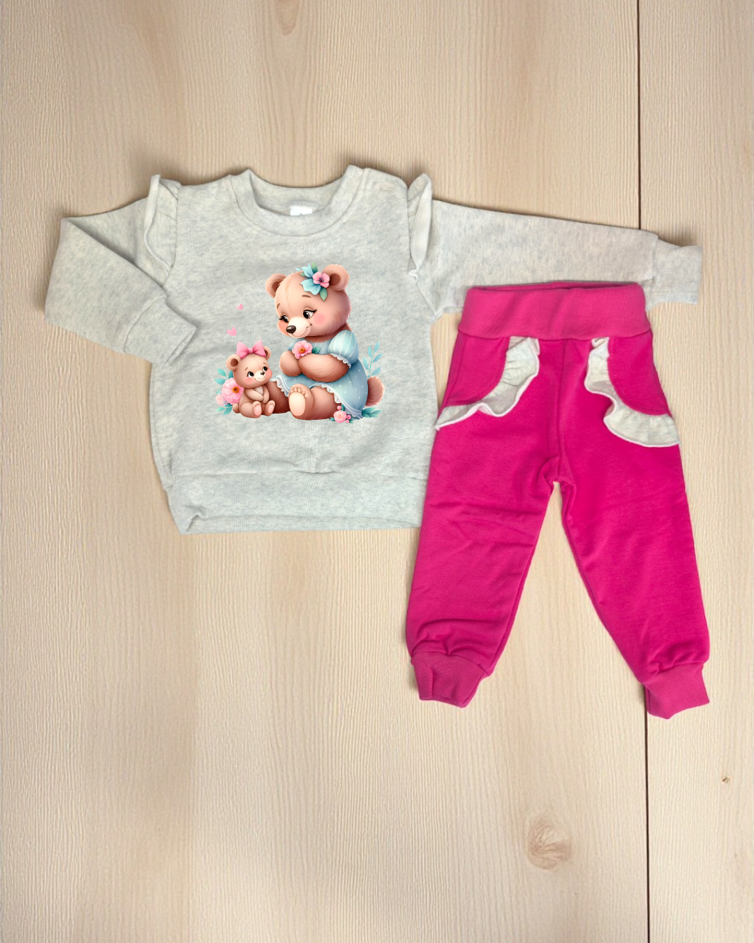 Conjunto de roupa infantil com camisola cinza estampada e calças rosas com bolsos cinza, sobre fundo de madeira clara.