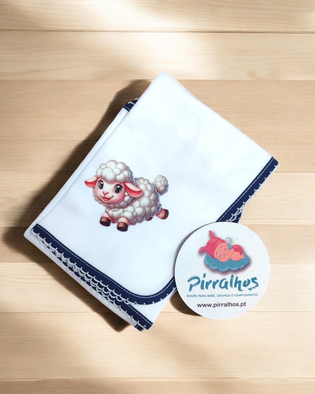 Fralda branca com borda evelho azul escura e bordado de ovelha sobre madeira clara.