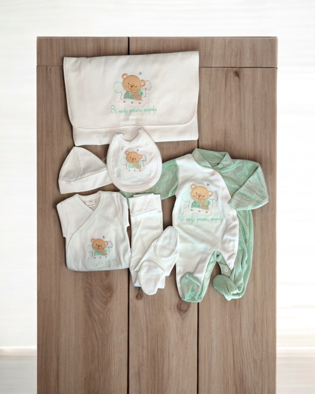 Conjunto de roupa de bebé branco e verde claro com ursinho e texto bordado, sobre mesa de madeira clara