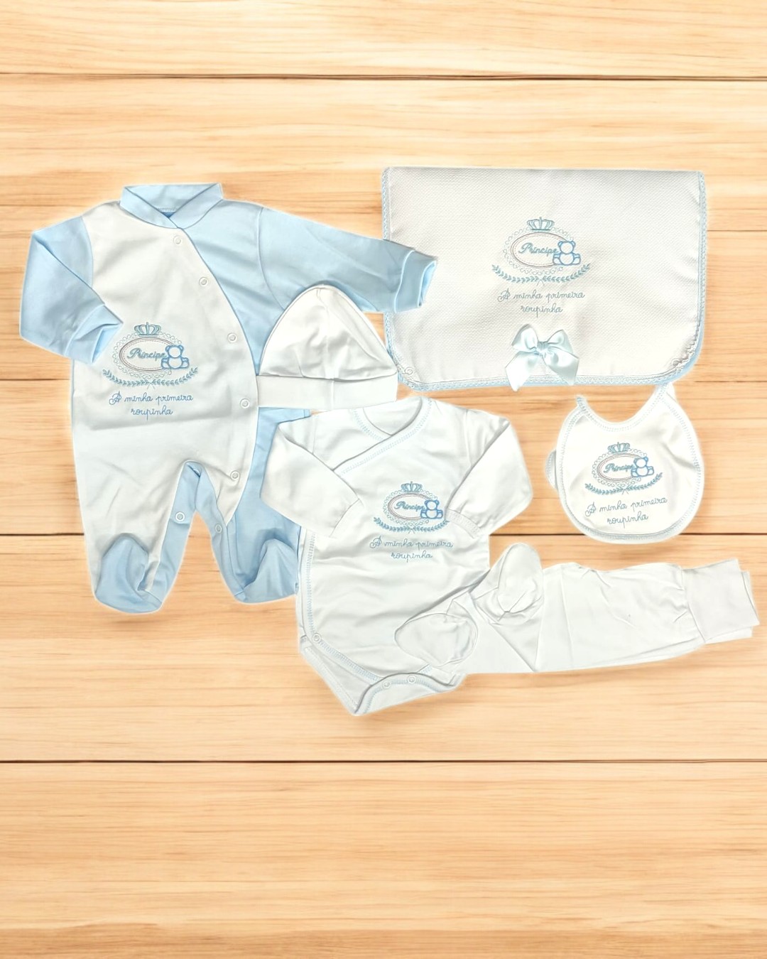 Conjunto de roupa para bebé azul claro e branco com bordados e manta sobre superfície de madeira clara
