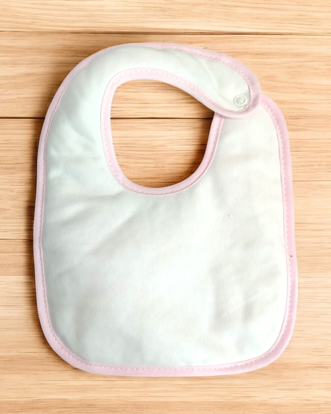 Babete branco com bordas rosa e fecho de pressão, sobre superfície de madeira clara