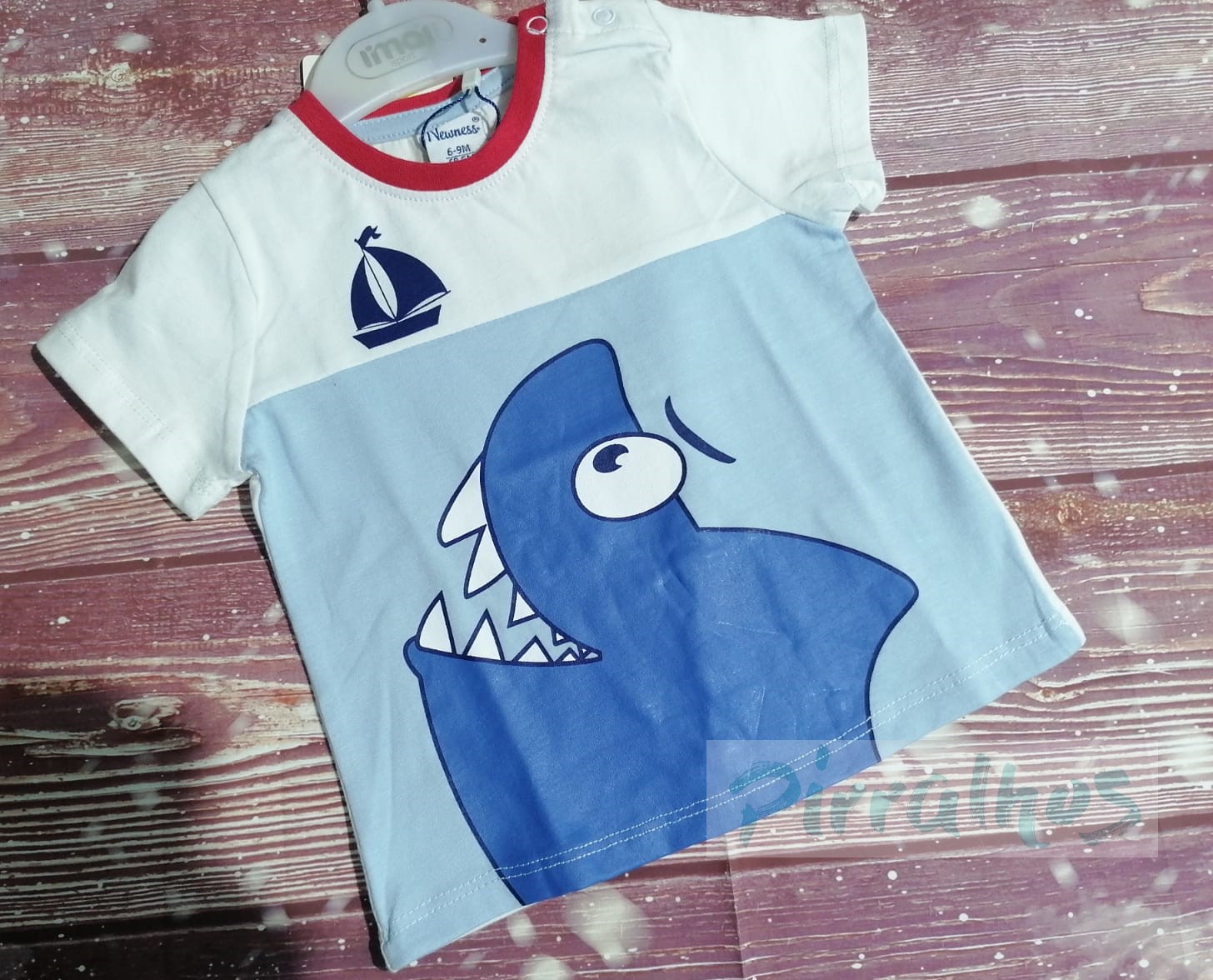 Camiseta infantil branca e azul com tubarão e vela, pendurada num cabide