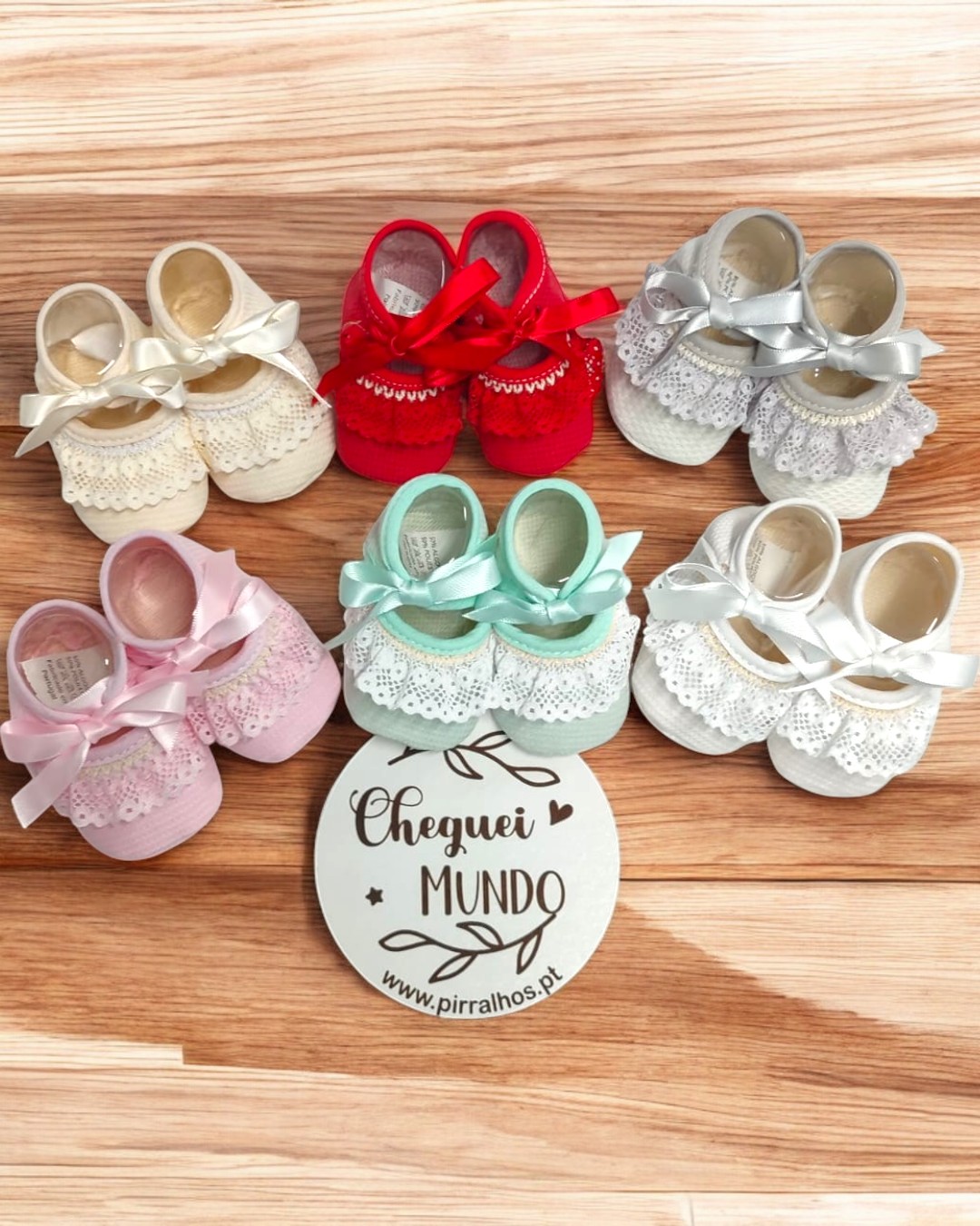 Seis pares de sapatinhos de bebé coloridos com laços e renda, dispostos em círculo com cartão central sobre madeira clara.