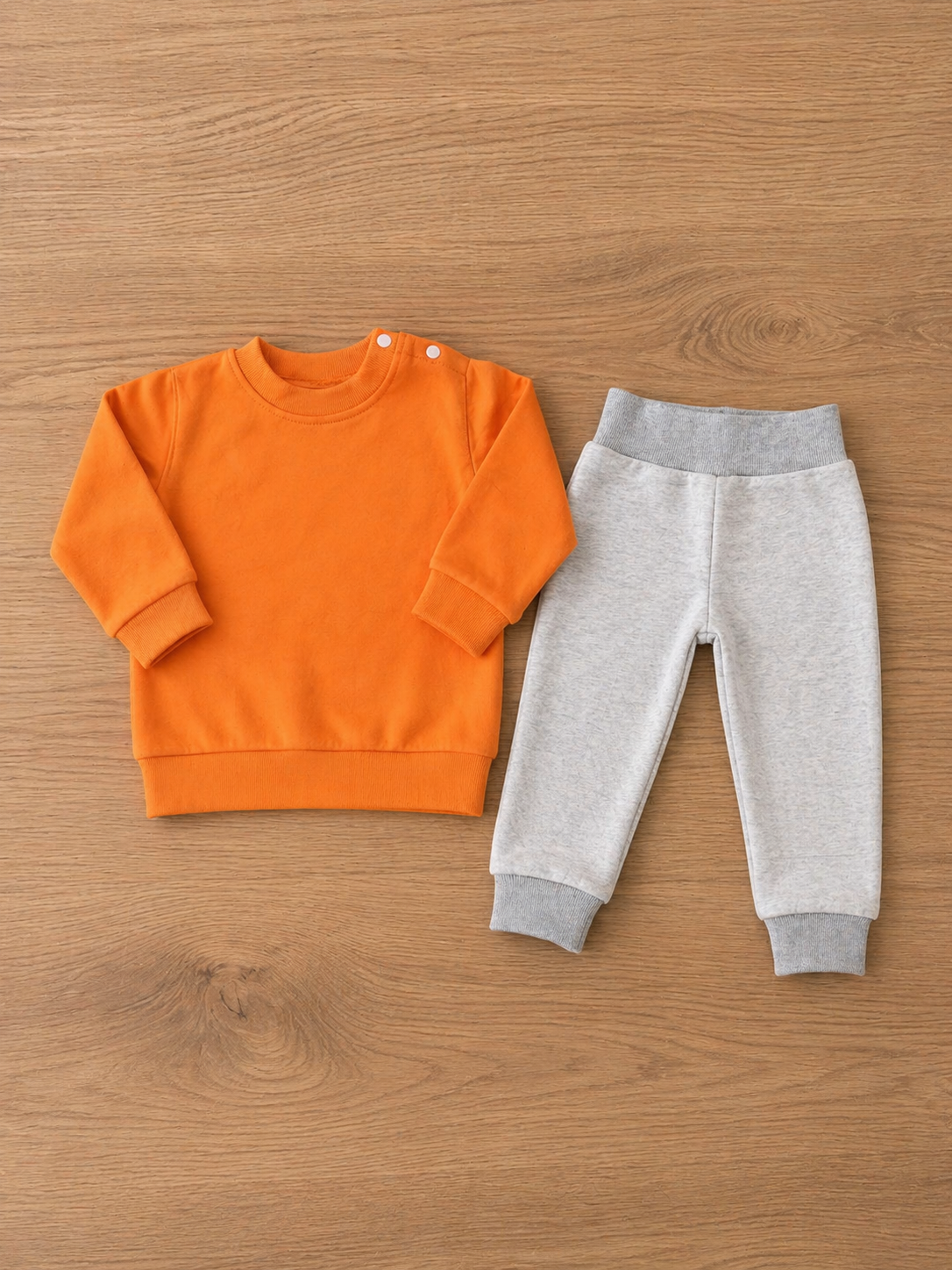 Conjunto de roupa de bebé com camisola laranja e calças cinza sobre madeira.