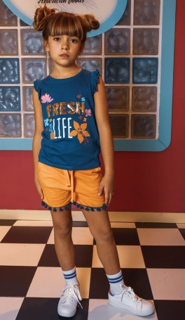 Menina com t-shirt azul e calções laranja em piso xadrez preto e branco com parede de vidro ao fundo