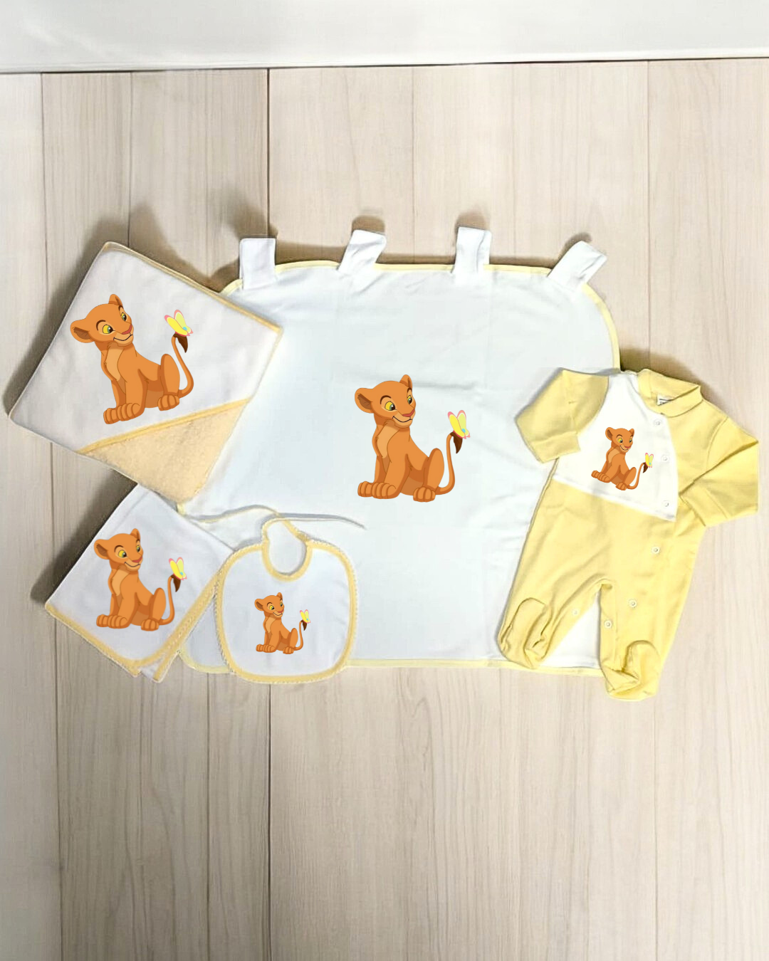 Conjunto de bebé amarelo e branco com estampas de leãozinho, incluindo babete, toalha, manta e pijama.