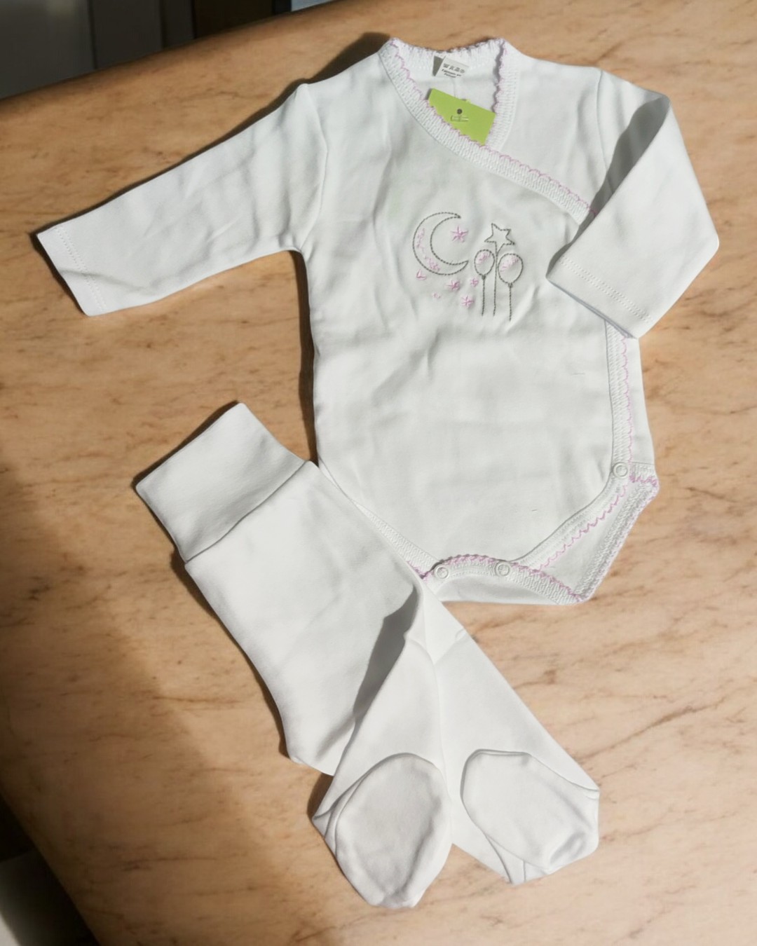 Conjunto de roupa de bebé branco com bordado e calças de pé sobre superfície marmoreada