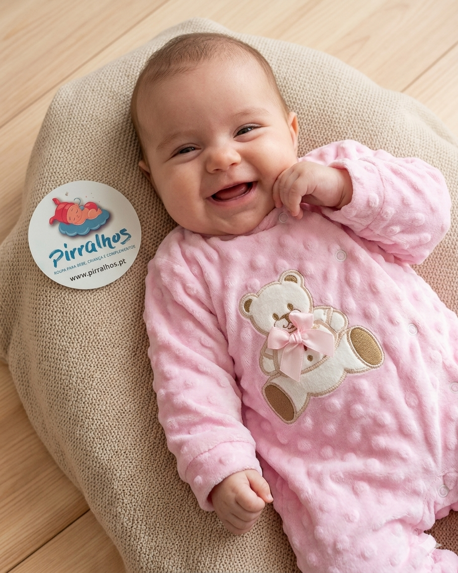 Bebé a sorrir com macacão rosa texturado e urso branco com laço cor-de-rosa deitado em almofada bege sobre chão de madeira