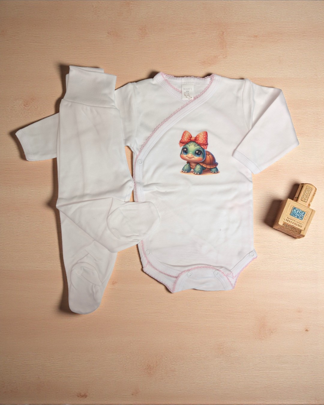 Conjunto de roupa de bebé branco com padrão de tartaruga e calças com pé em superfície de madeira clara