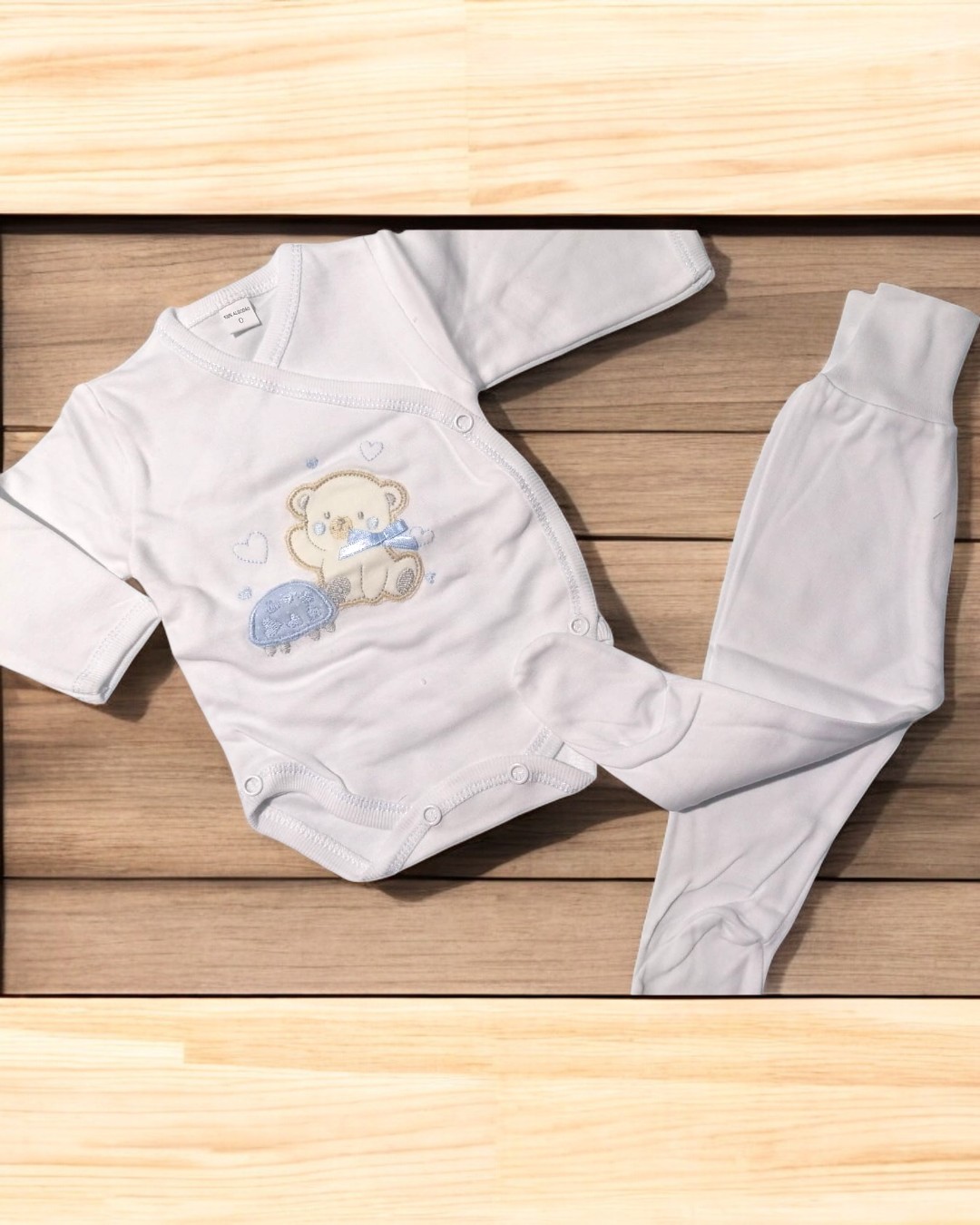 Conjunto de roupa de bebé branco com ursinho e carrinho azul sobre madeira clara