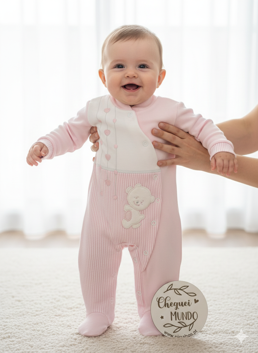 Bebé a usar macacão rosa e branco com aplicação de urso e corações, segurado por mãos.