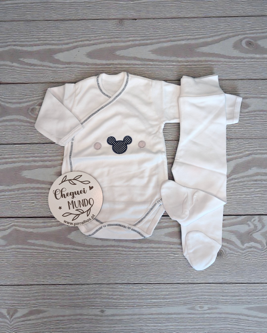 Conjunto de bebé branco com padrão e figura de orelhas pretas sobre fundo de madeira cinza.