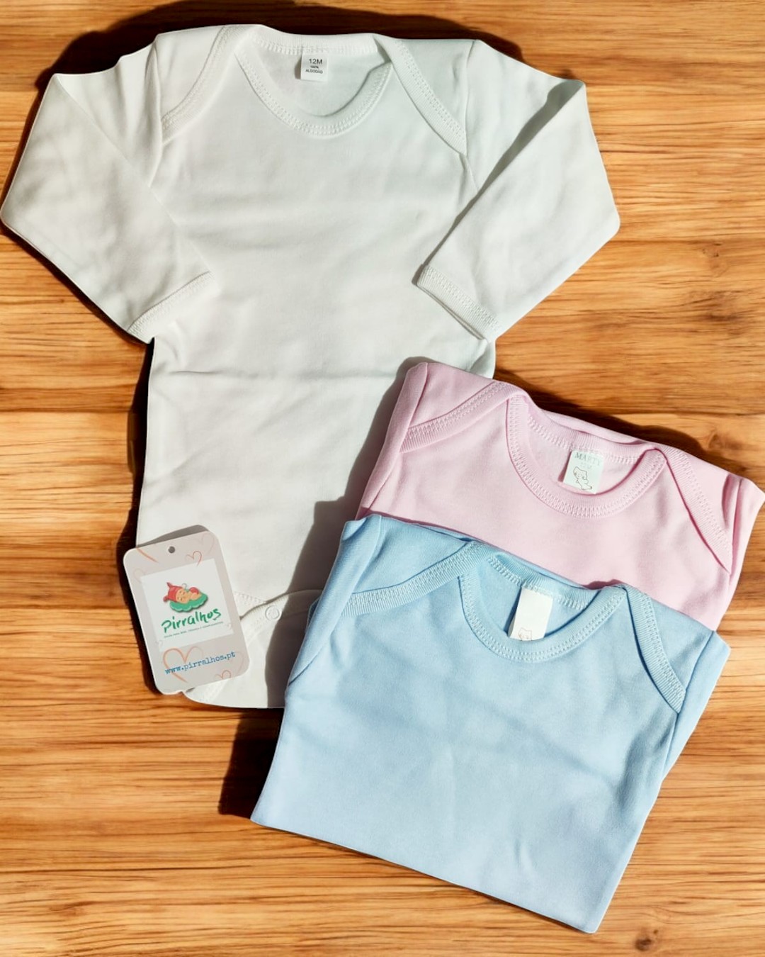Três bodies de bebé em branco, rosa e azul claro com etiquetas com o texto Pirulinhos, sobre madeira clara.