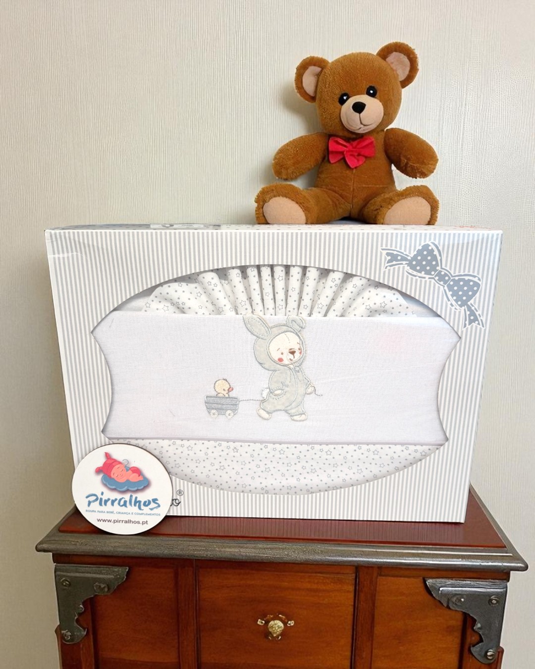 Conjunto roupa bebé branco com coelhinho bordado, em caixa decorada, sobre móvel de madeira com ursinho de peluche castanho.