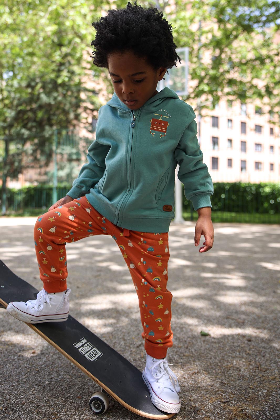 Criança com casaco azul, calças laranjas estampadas e ténis brancos num skate