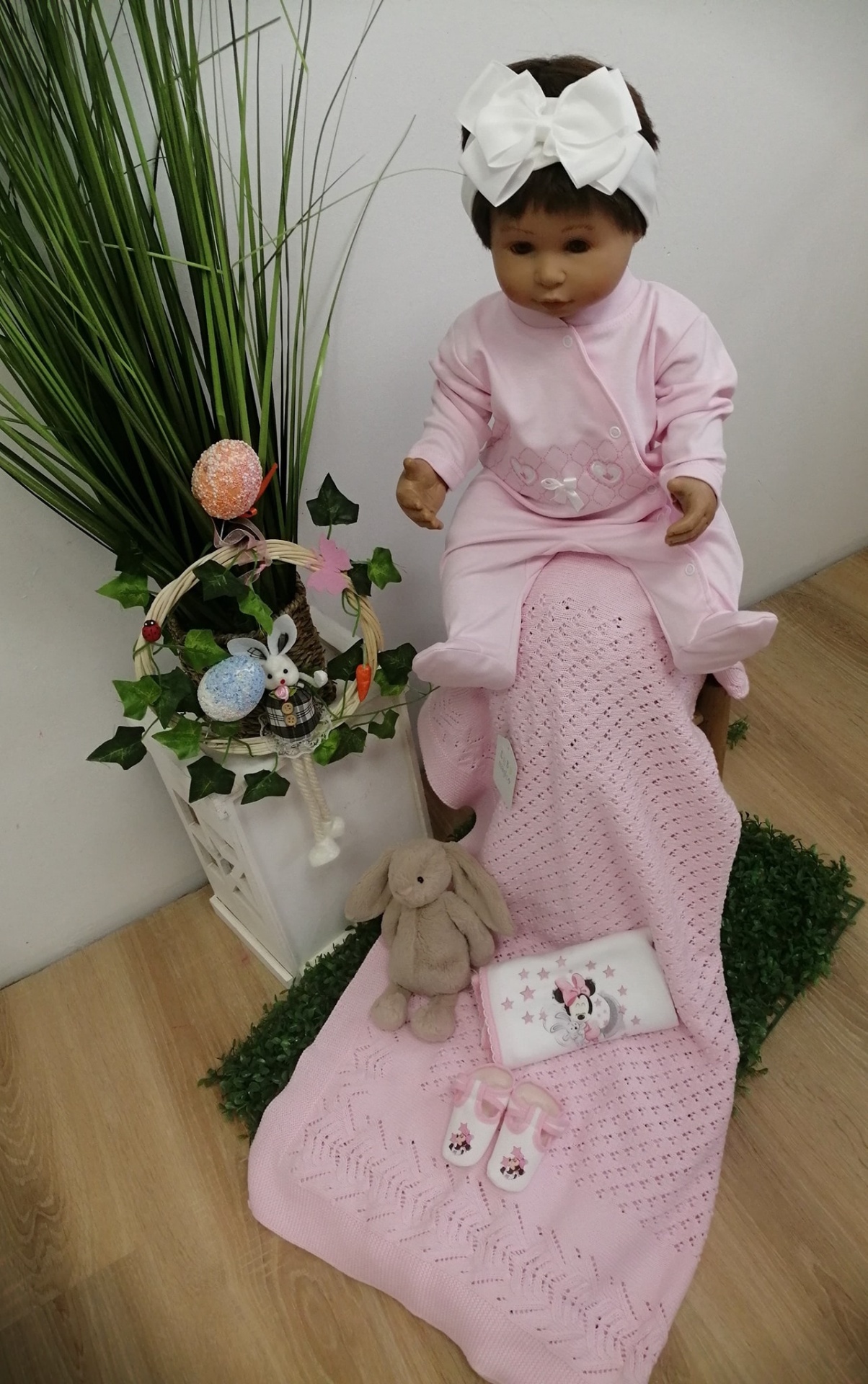 Boneca bebé rosa com faixa branca sentada em cobertor rosa com brinquedo coelho e acessórios