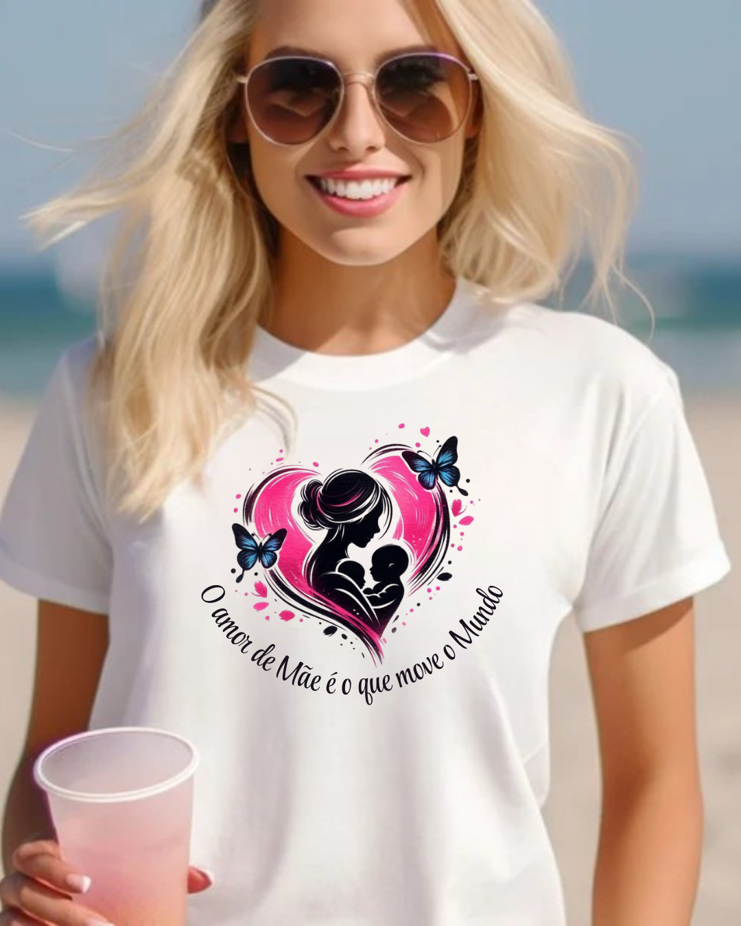 T-shirt branca com estampa de mãe e bebé em coração rosa e texto em português, usada por mulher na praia