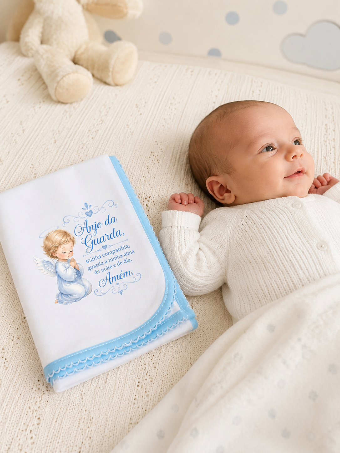 Manta branca e azul com ilustração de anjo e texto, bebé deitado em cama bege com padrão de nuvens