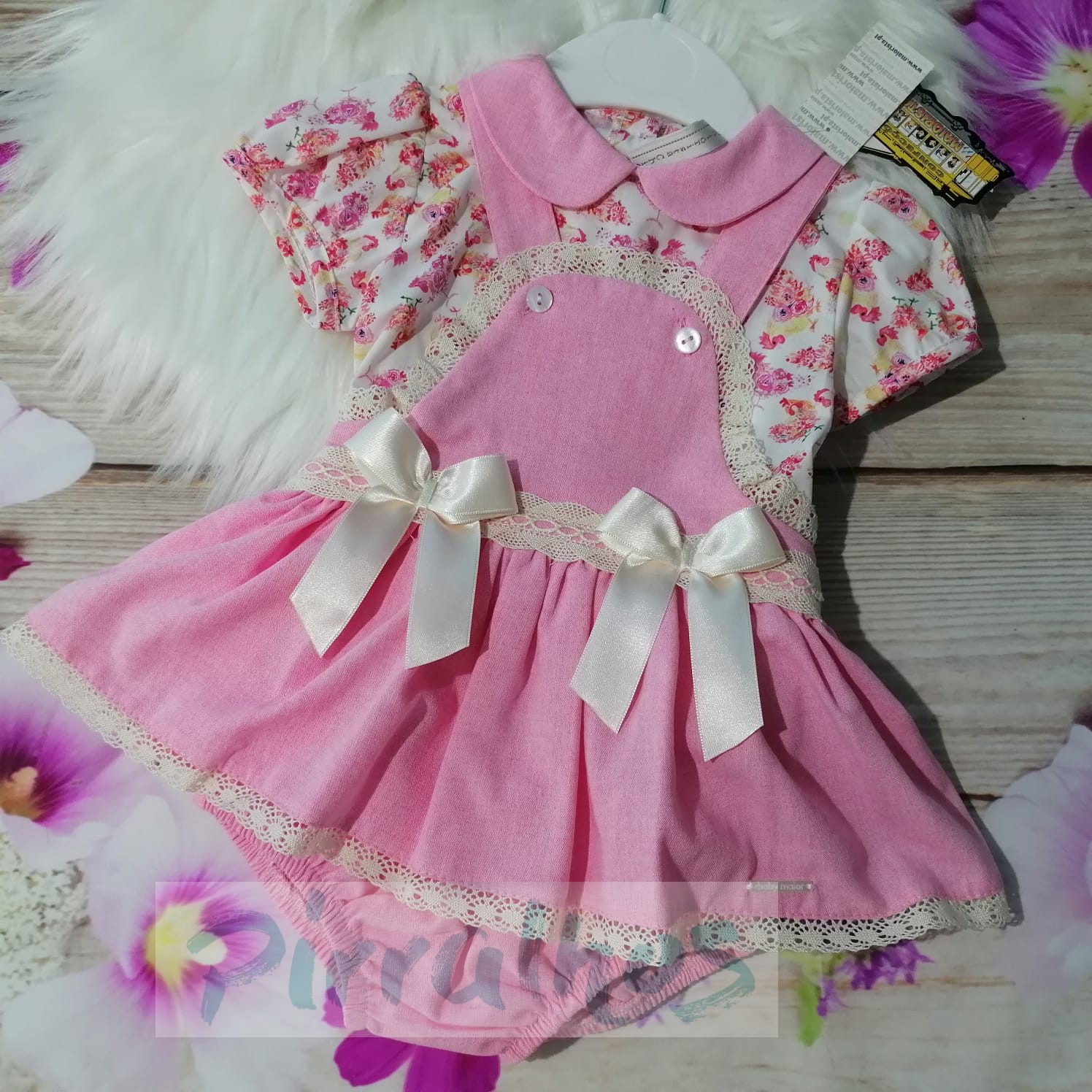 Vestido rosa para bebé com padrão floral, laços e detalhes em renda creme numa superfície de madeira com flores artificiais.