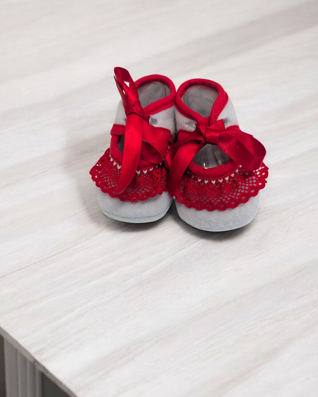 Sapatinhos de bebé cinza claro com renda e fitas vermelhas sobre mesa clara