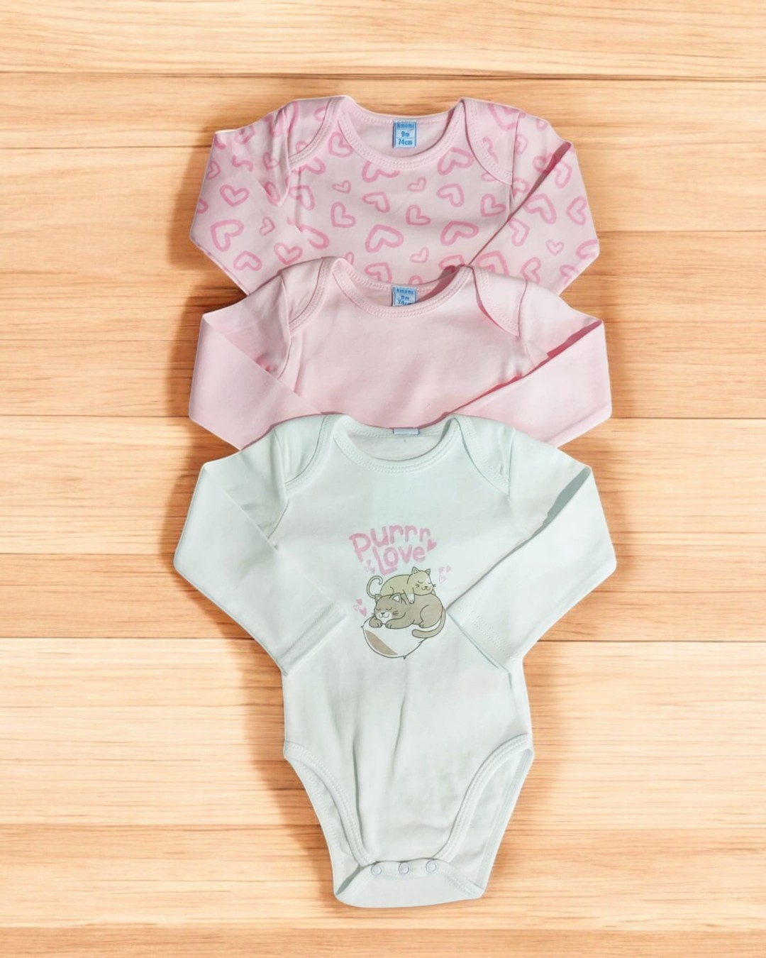 Três bodies de manga comprida para bebé em branco com estampa de gato e texto, rosa liso e rosa com corações, sobre superfície de madeira.