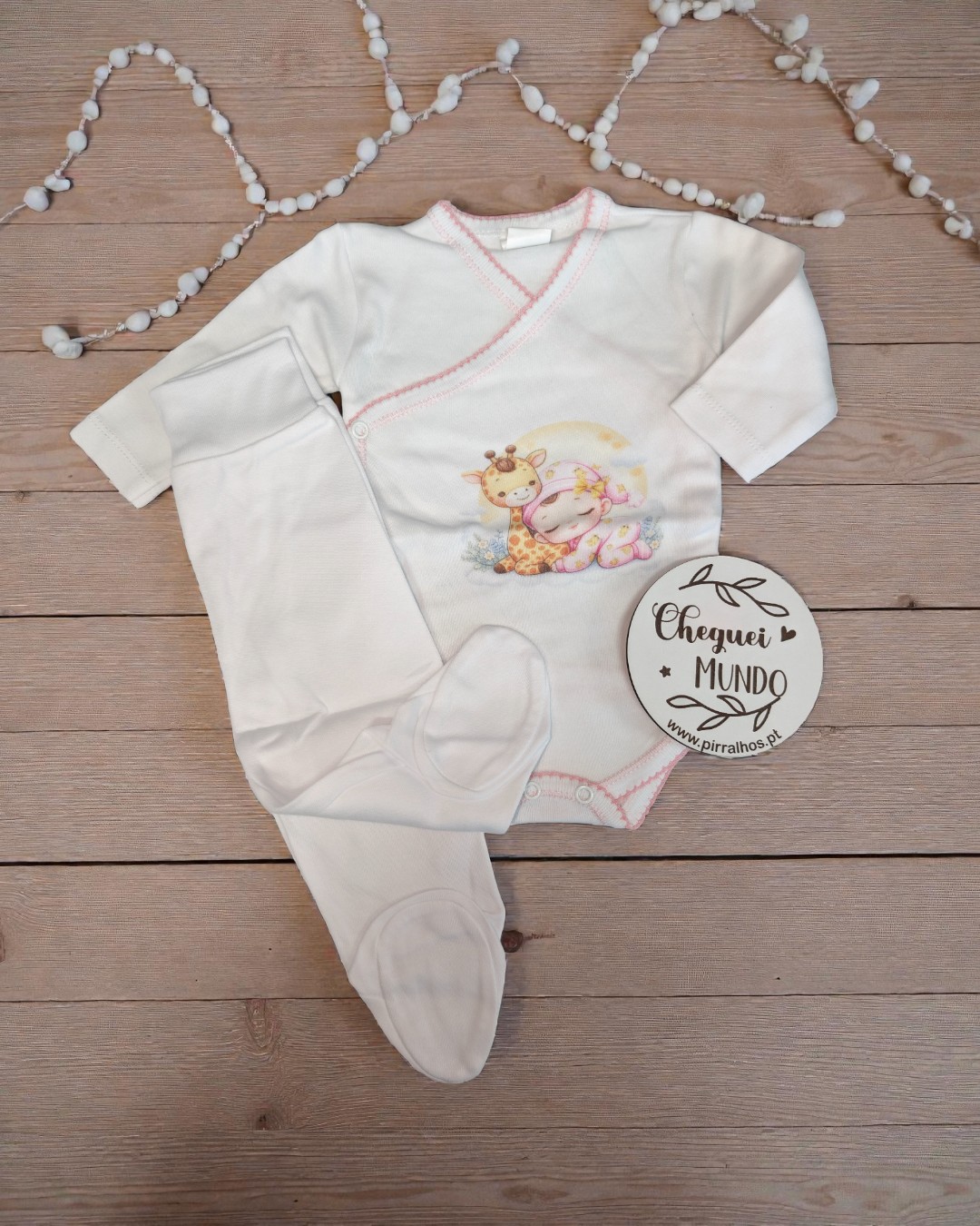 Conjunto de roupa de bebé branco com estampa de girafa e bebé, etiqueta com texto, decorado com guirlanda branca e fundo de madeira.