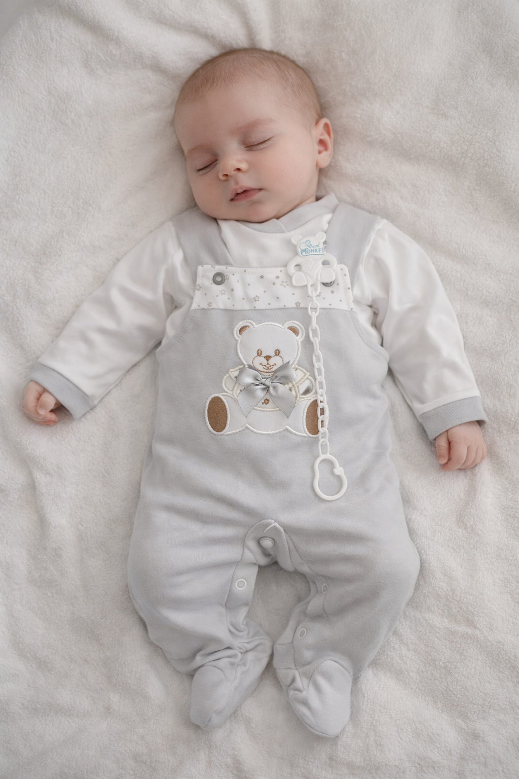 Bebé com macacão cinzento com estampa de urso deitado numa manta branca