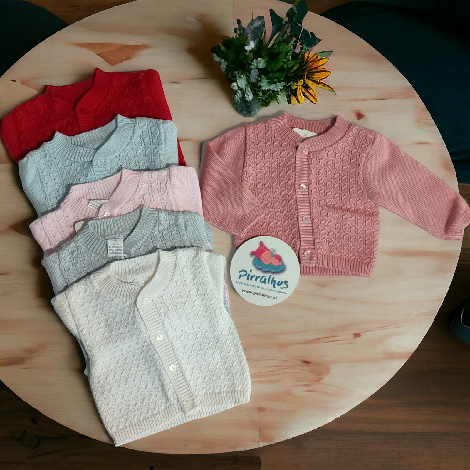 Casacos de malha para bebés coloridos dispostos sobre mesa redonda de madeira clara com etiqueta Pirralhos e vaso de flores