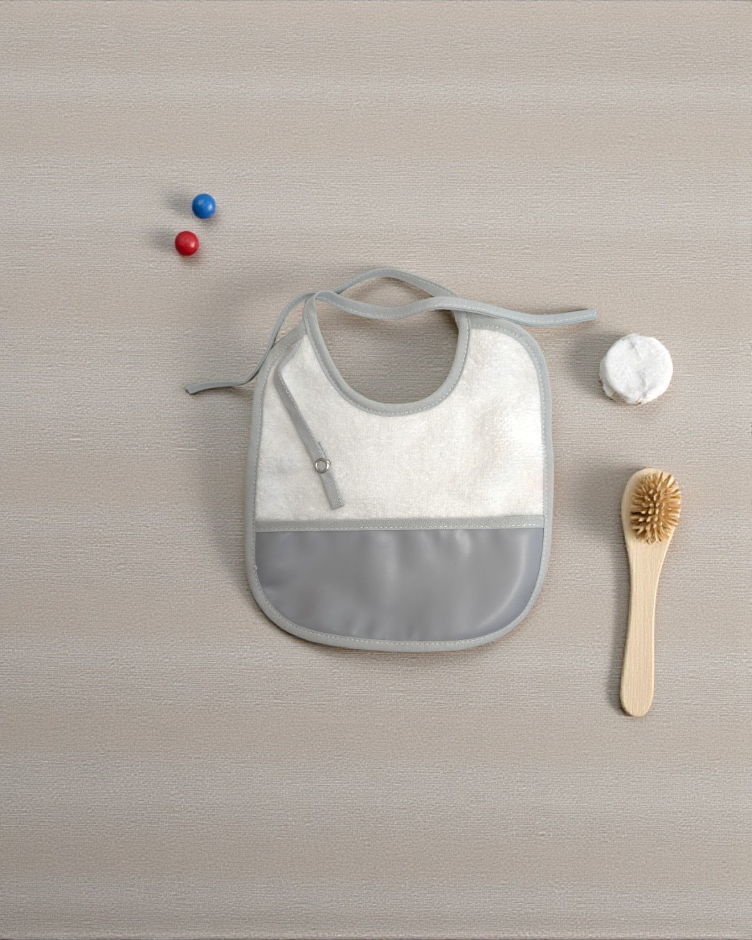 Babete branco com bolso cinzento e fita para atar rodeado por escova de madeira, disco de algodão e bolas azul e vermelha numa superfície bege.