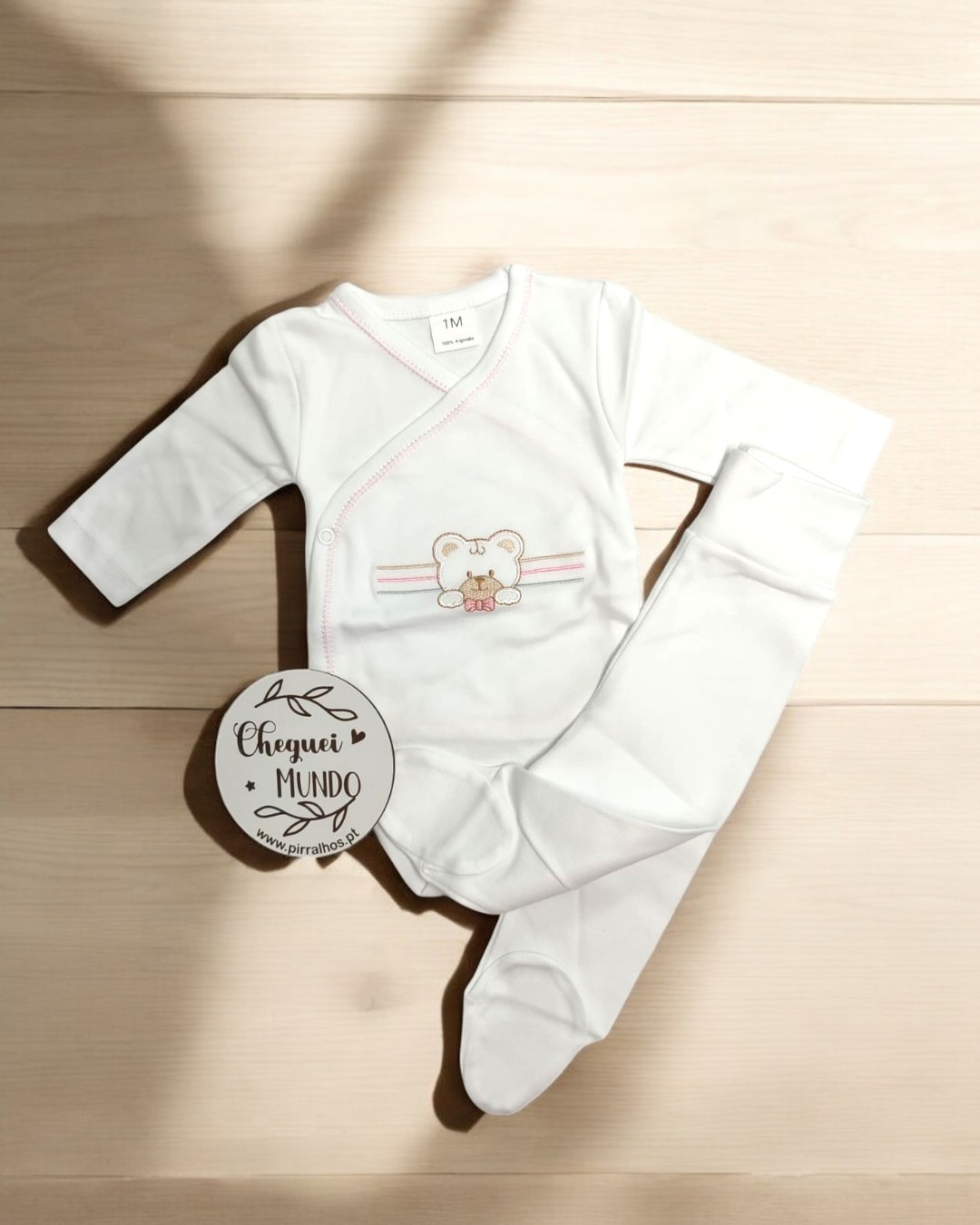 Pijama branco de bebé com ursinho bordado e riscas cor-de-rosa sobre chão de madeira clara