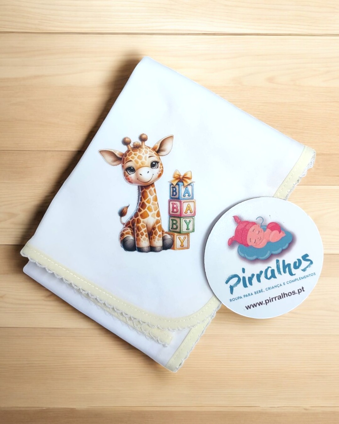 Fralda branca com girafa e blocos de letras, borda em croché creme, sobre madeira clara com adesivo da marca Pirralhos