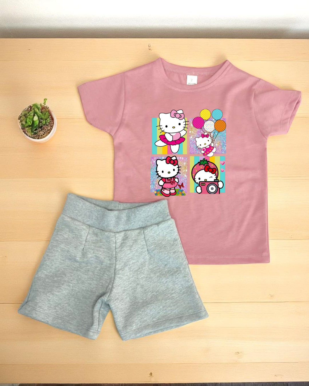 Conjunto infantil rosa Hello Kitty e calções cinza sobre madeira clara com planta ao lado.