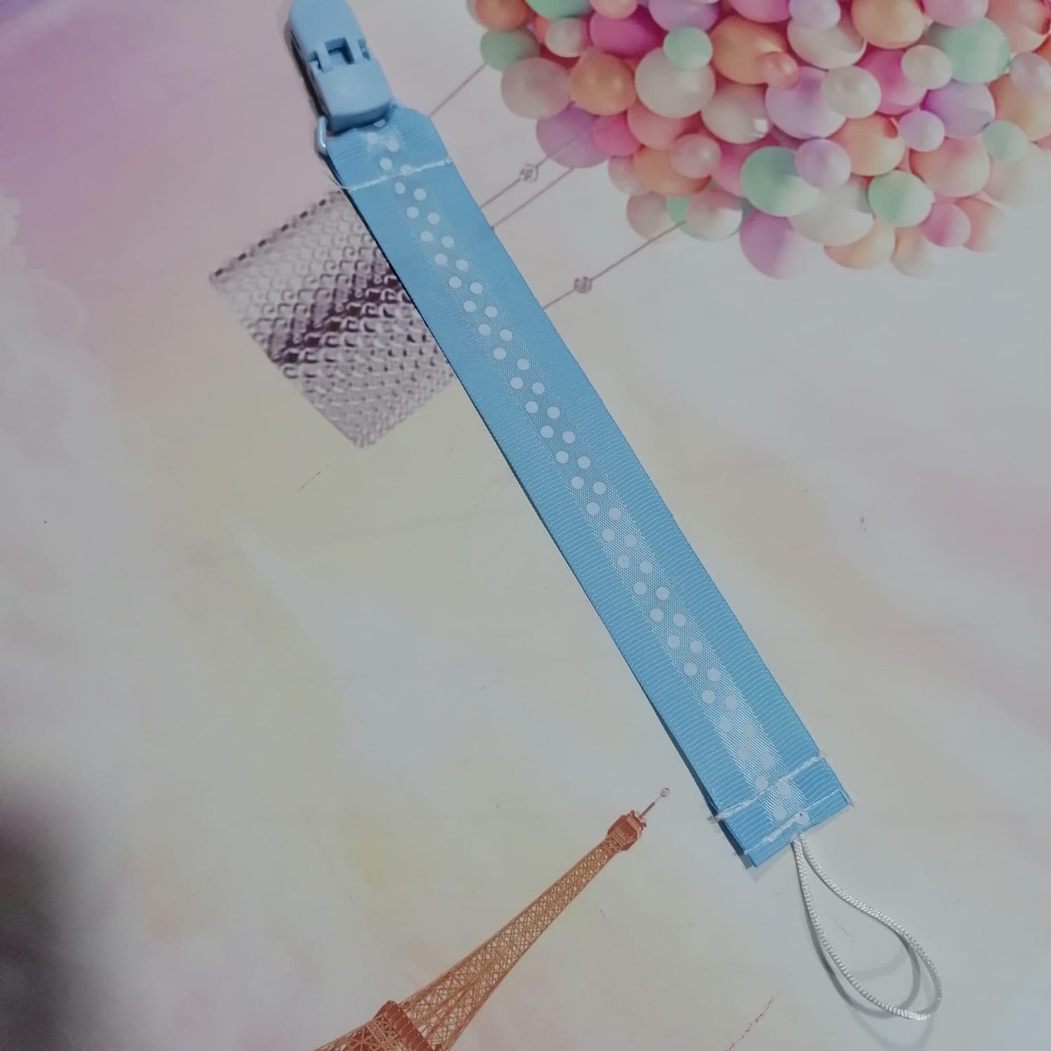 Pulseira azul claro de tecido com padrão de bolinhas e fecho de plástico em fundo com Torre Eiffel e balão colorido