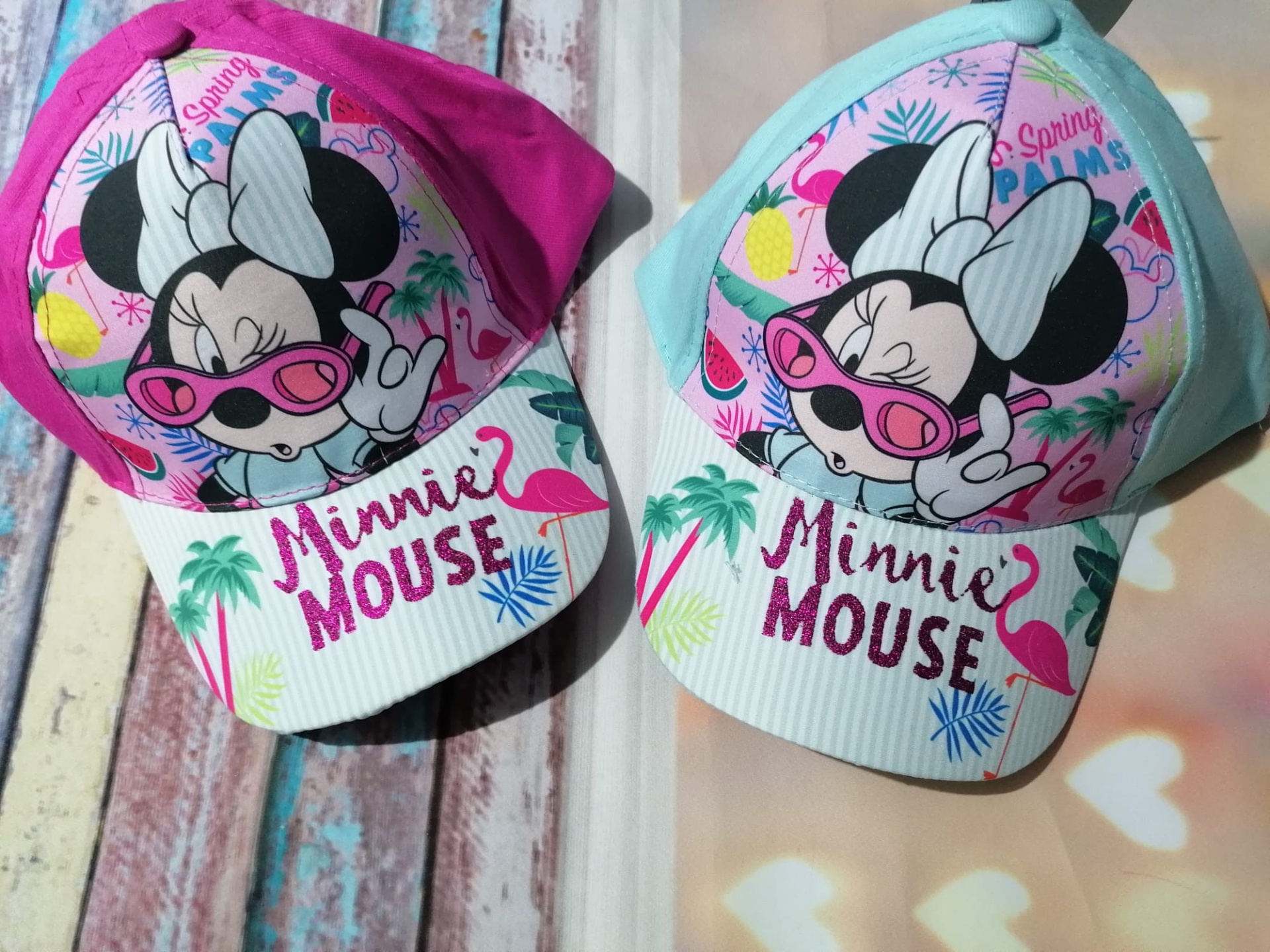 Dois bonés infantis da Minnie Mouse, um rosa e outro azul claro, com desenho e texto colorido.