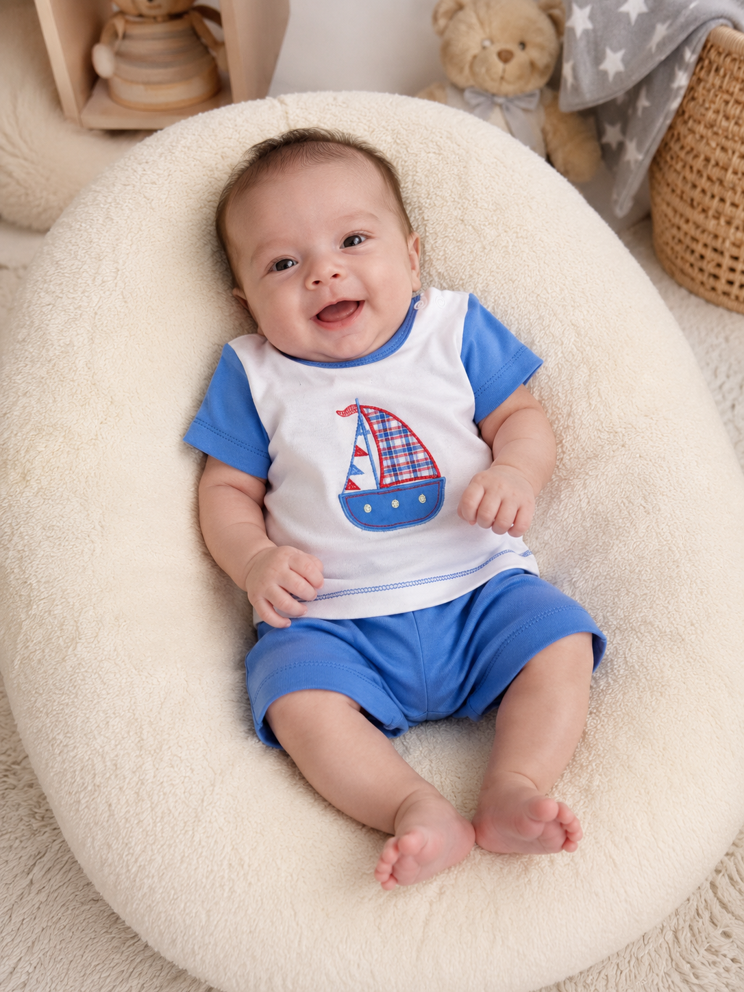 Bebé sorridente com conjunto branco e azul com barco, sentado em almofadão creme felpudo.
