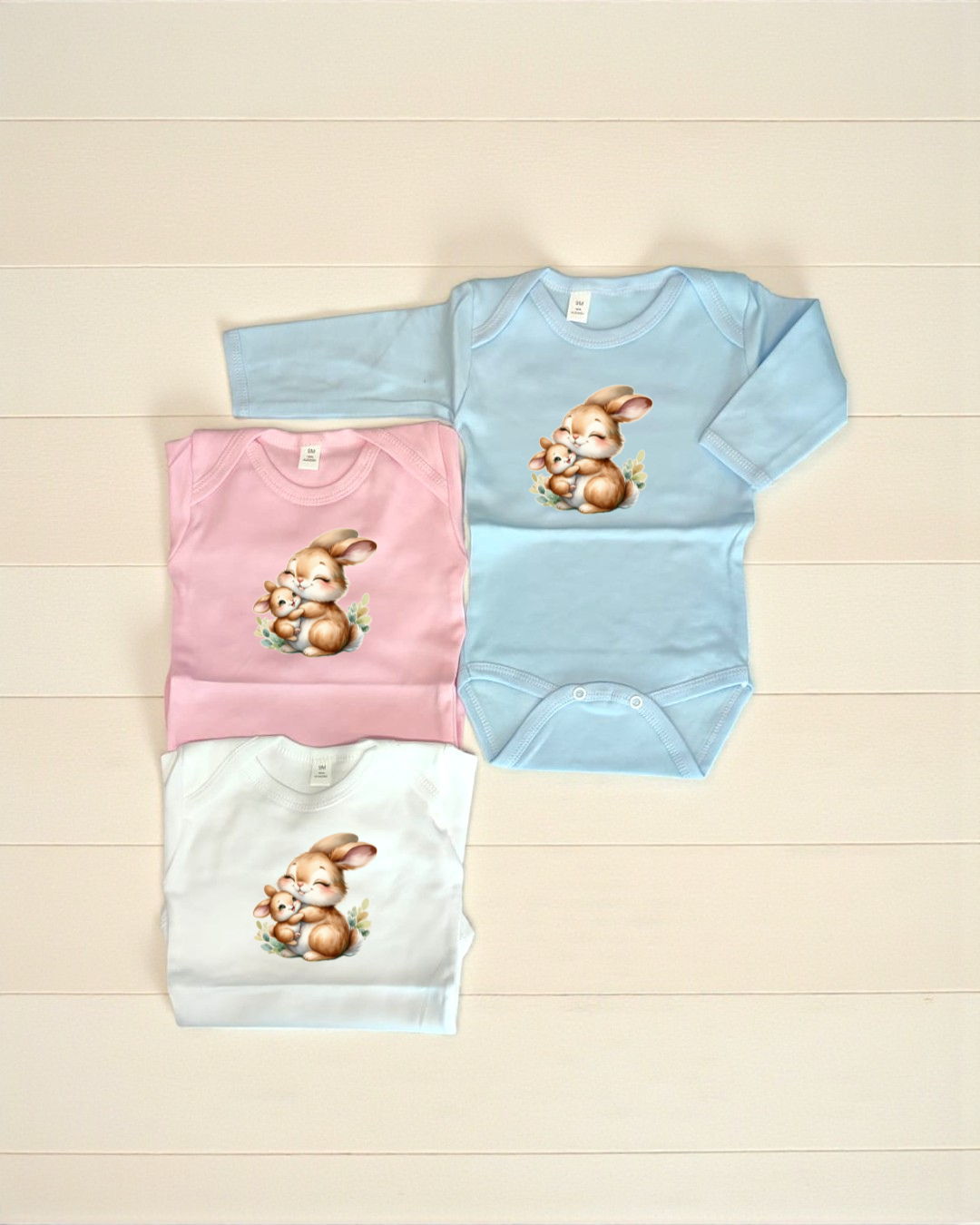 Três bodies de bebé com desenho de lebres abraçadas em cores azul, rosa e branco sobre fundo branco.