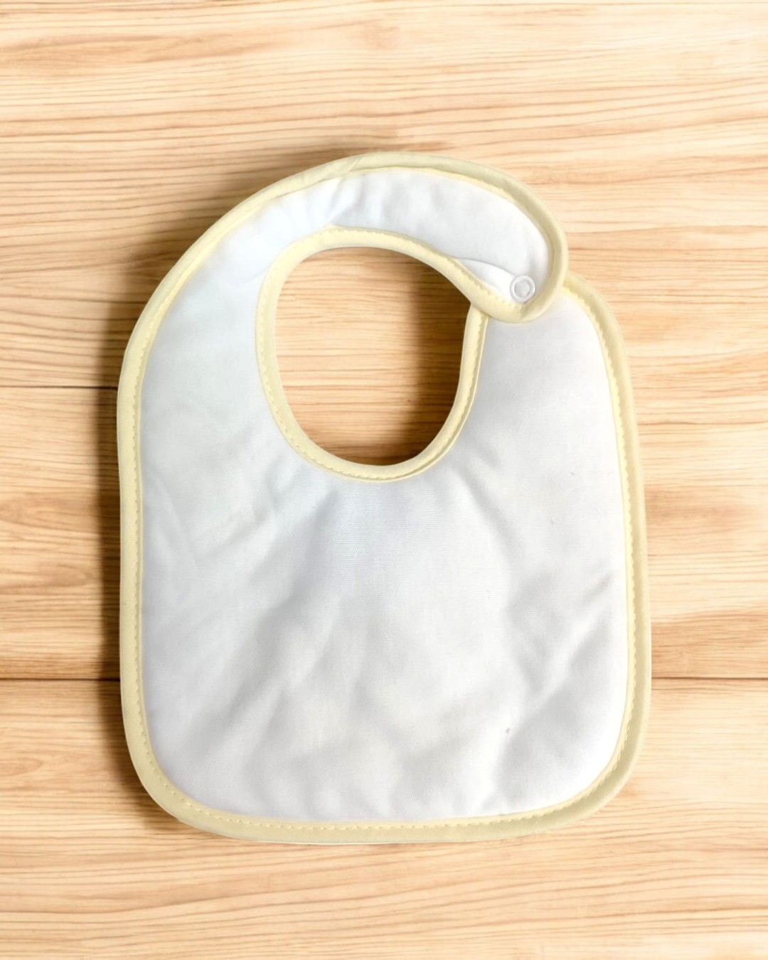 Babete branco para bebé com borde amarelo claro e fecho de pressão sobre fundo de madeira