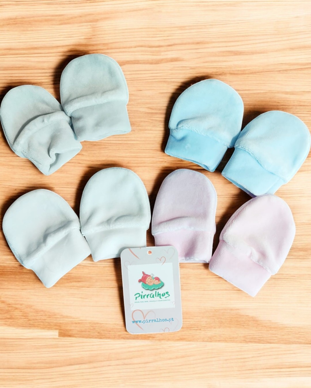 Luvas para bebé em várias cores claras com etiqueta Pirilahes em fundo madeira