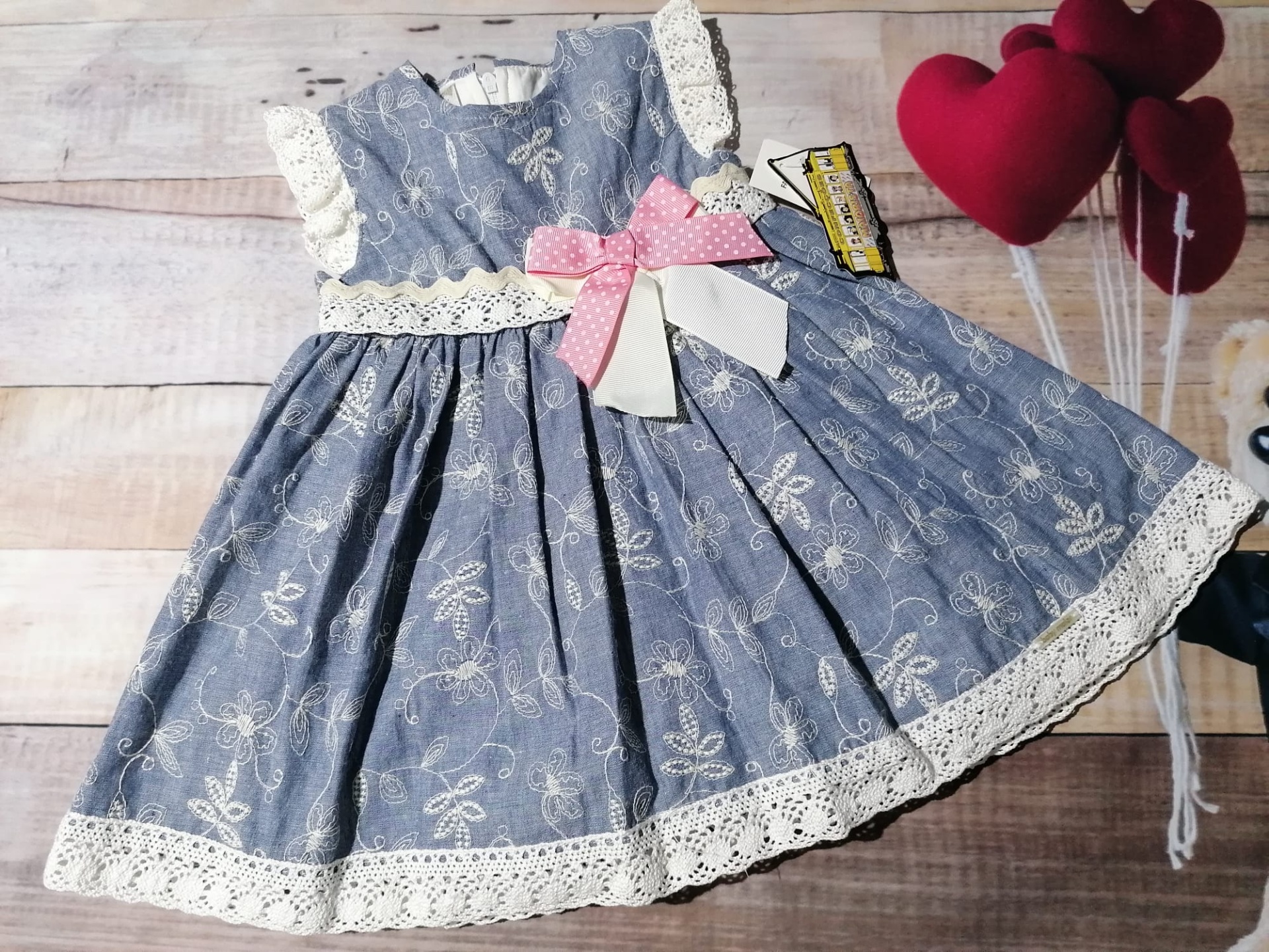 Vestido infantil azul com padrão floral branco e renda branca, laço rosa e branco, etiqueta e balões de coração vermelhos