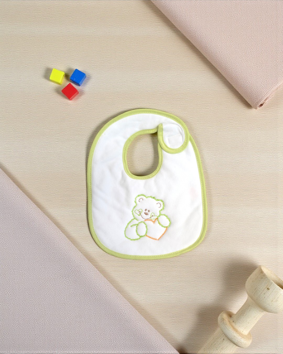 Babete branco com borda verde claro e bordado de ursinho colorido sobre superfície clara ao lado de cubos coloridos