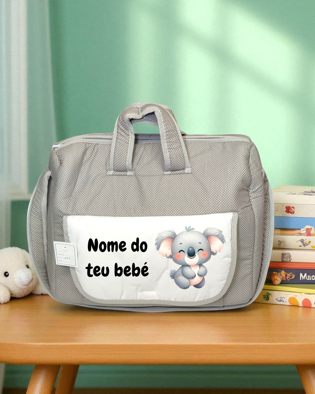Bolsa cinzenta para bebé com bolso branco e ilustração de coala, junto a livros infantis e peluche.