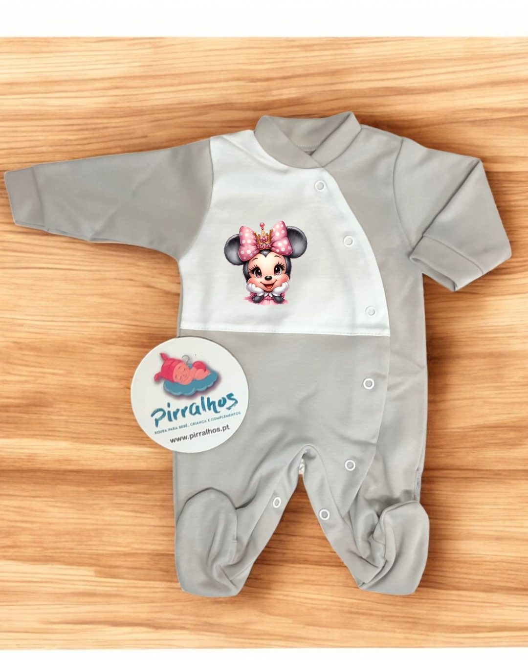 Pijama de bebê cinza e branco com estampa de personagem Minnie com coroa e etiqueta Pirralhos sobre madeira clara.