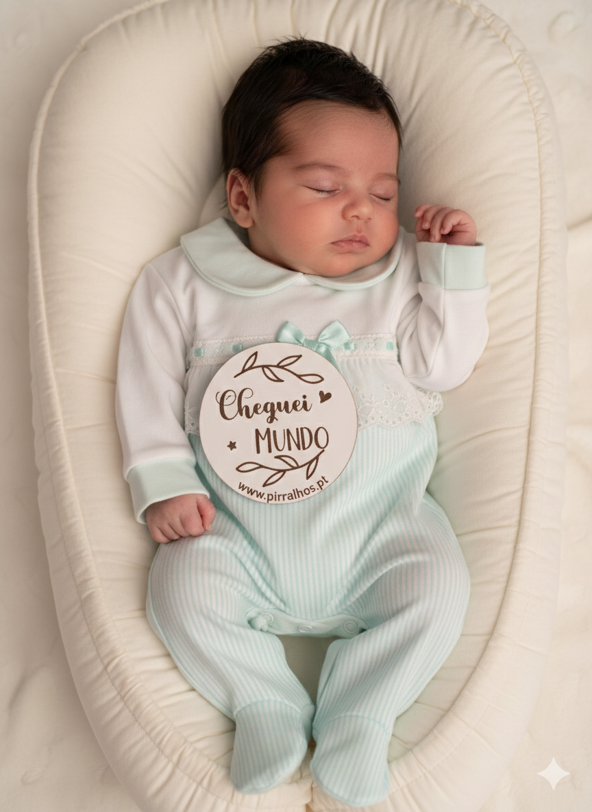 Bebé a dormir em almofada branca com macacão verde claro às riscas e branco com colarinho e laço, segurando placa de madeira com texto