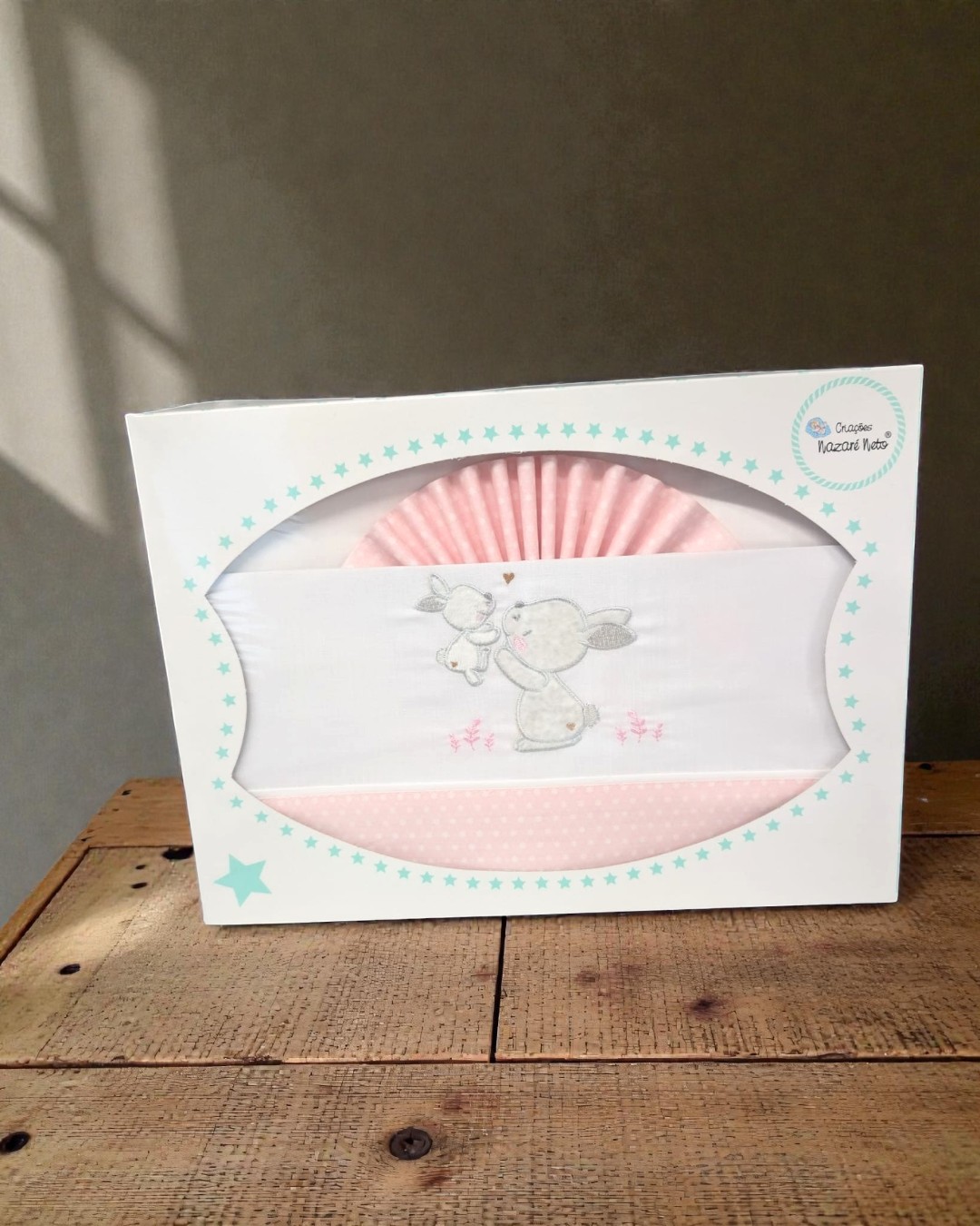 Conjunto de roupa de bebê rosada e branca com bordado de coelhos em caixa branca com estrelas azuis