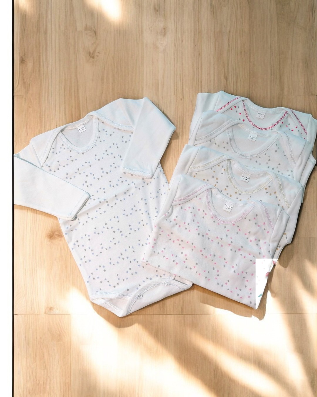 Bodies de bebé brancos com padrão de pontos coloridos em cruz sobre chão de madeira clara