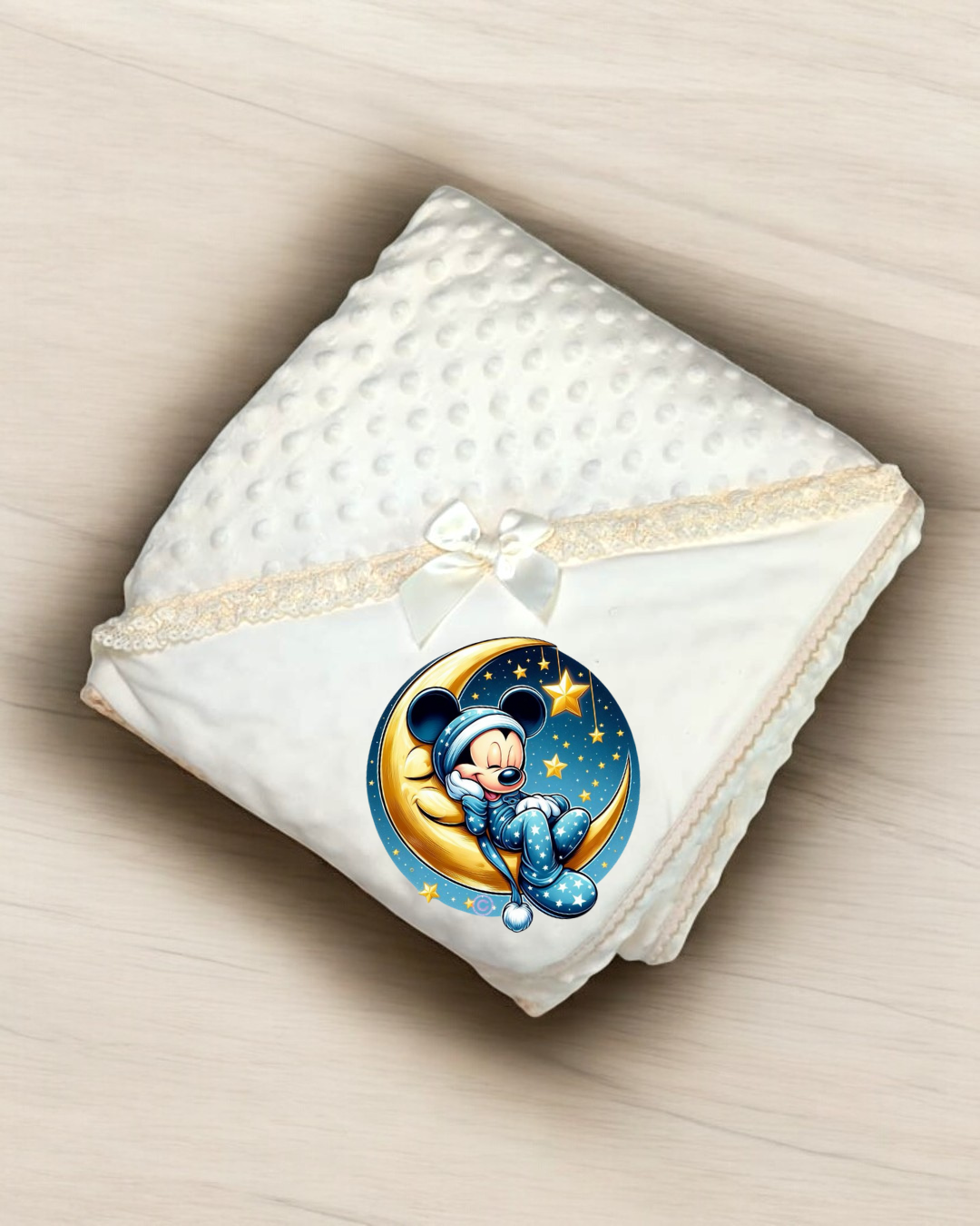 Manta branca com textura bolinha e borda de renda creme, com laço branco e imagem de Mickey Mouse em pijama azul sentado numa lua sobre fundo de estrelas.