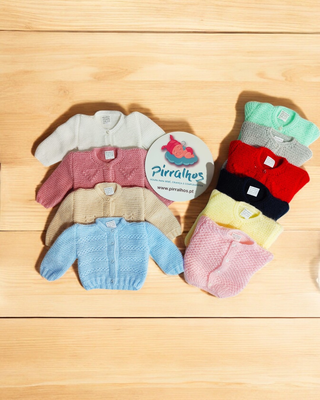 Casacos de bebé coloridos em malha sobre madeira com cartão redondo 'Pirralhos www.pirralhos.pt'.