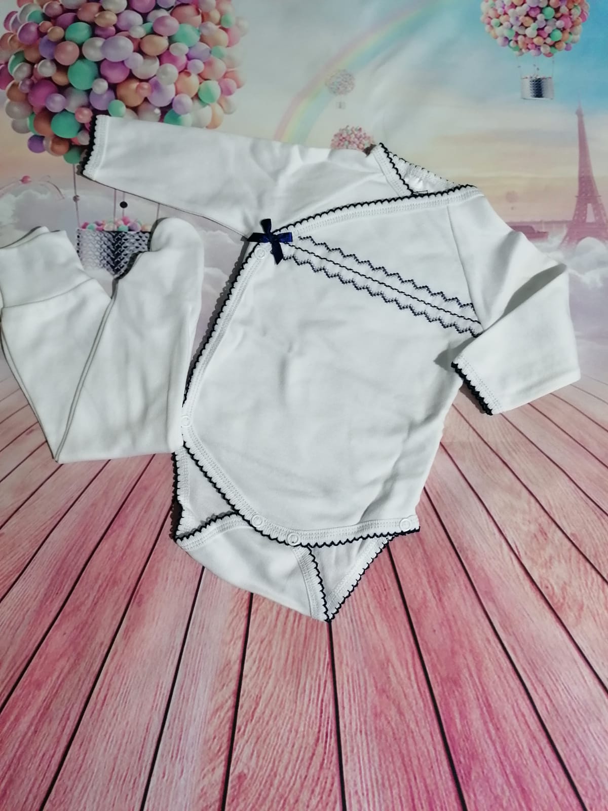 Roupa de bebé branca com detalhes pretos num fundo com Torre Eiffel e balões