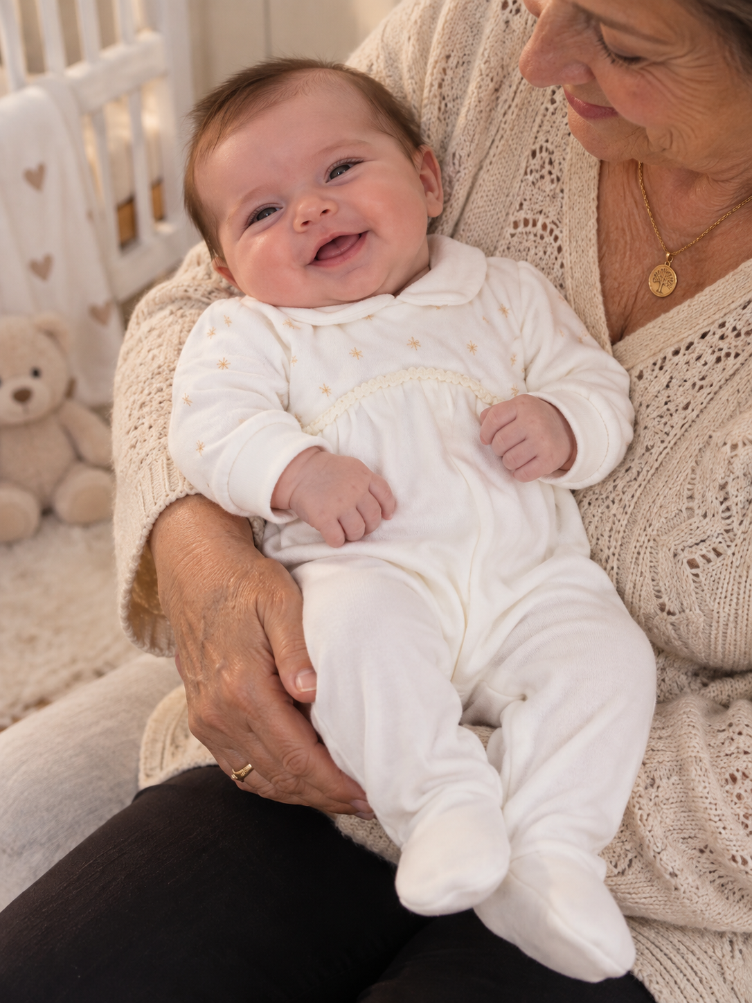 Bebé sorridente com babygrow branco com estrelas douradas nas mãos de mulher com camisola bege de malha.