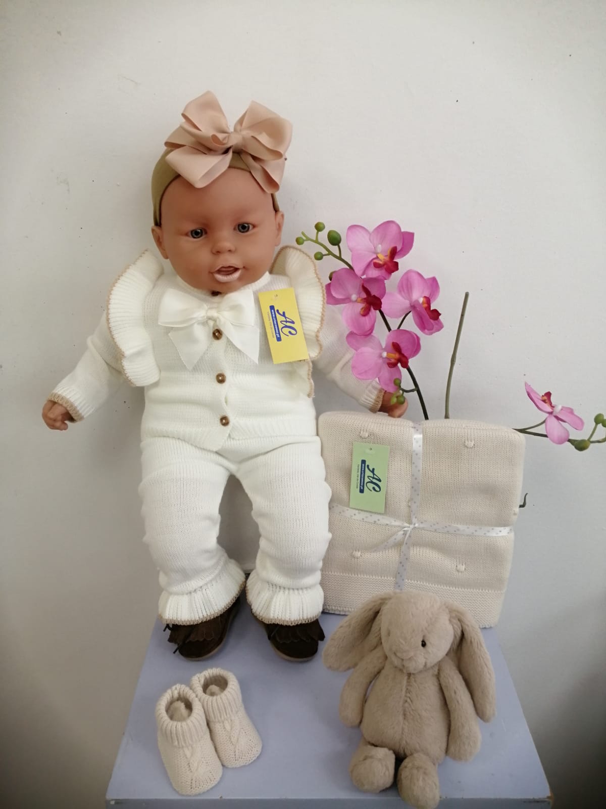 Boneca bebé de malha branca com laço rosa, manta, botinhas e coelho de peluche junto a uma orquídea rosa.